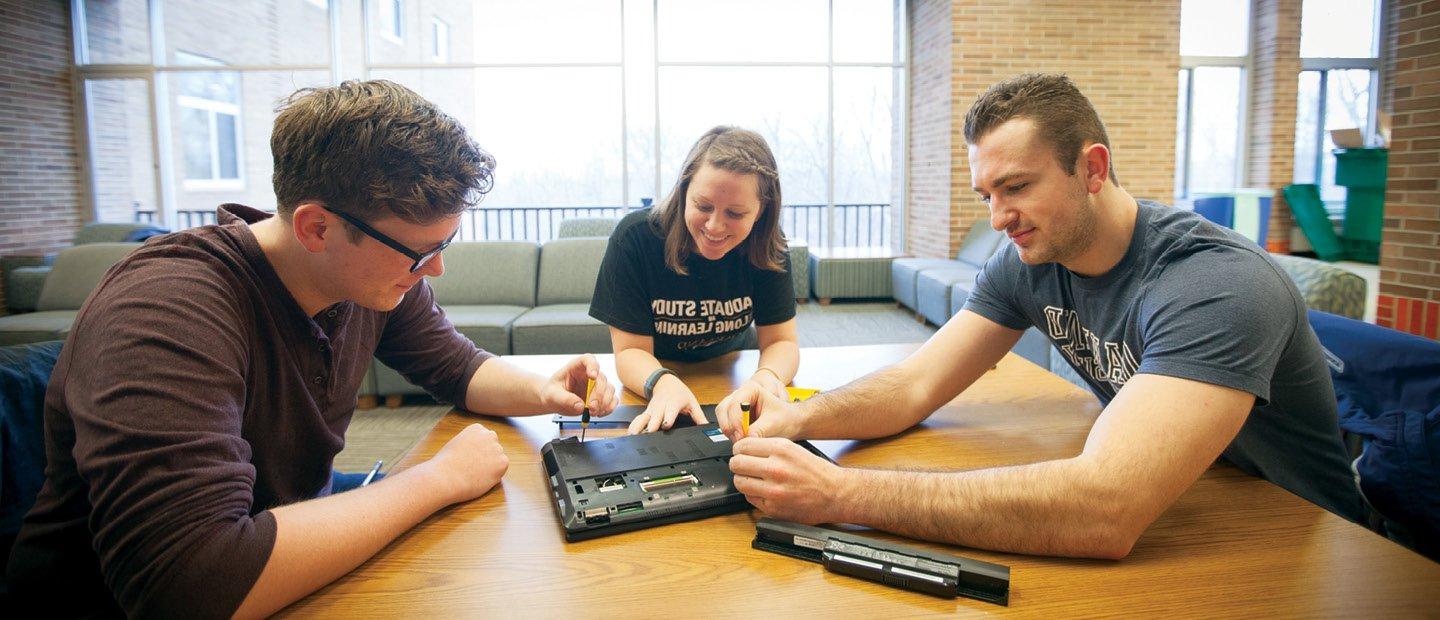three students seated around a table, working with screwdrivers to examine a laptop